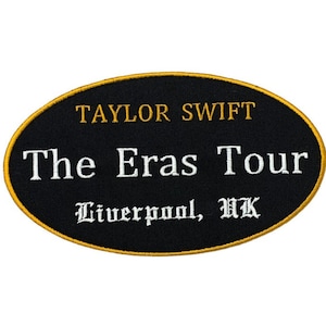 Peut inclure: Écusson ovale noir avec bordure dorée. Il affiche le texte "TAYLOR SWIFT" au-dessus de "The Eras Tour" et "Liverpool, UK" en blanc. Probablement un souvenir de concert.