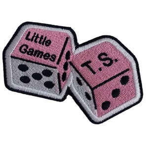 Puede incluir: Parche bordado con dos dados rosas con puntos negros y bordes blancos. Un dado dice "Little Games" y el otro "T.S." sobre un fondo blanco. El parche está diseñado para ropa o accesorios.
