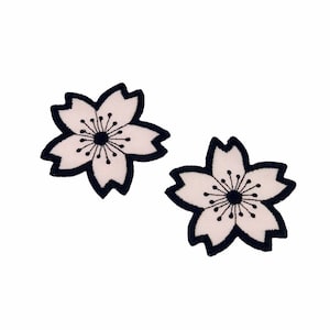 May include: Two embroidered flower patches with a white center, black outline, and black center detail. The patches are in the shape of a cherry blossom, with detailed stitching. The patches are on a white background.