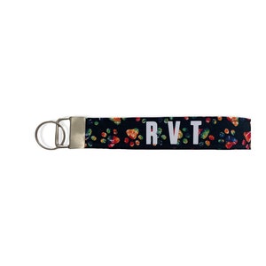 May include: Black wrist strap keychain with a colourful paw print pattern. The keychain has the letters "RVT" in white block letters. It features a silver metal clasp and key ring.