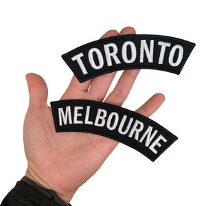 May include: Two black embroidered patches with white text. The top patch reads "TORONTO" and the bottom patch reads "MELBOURNE". Both patches are curved and have a black border.