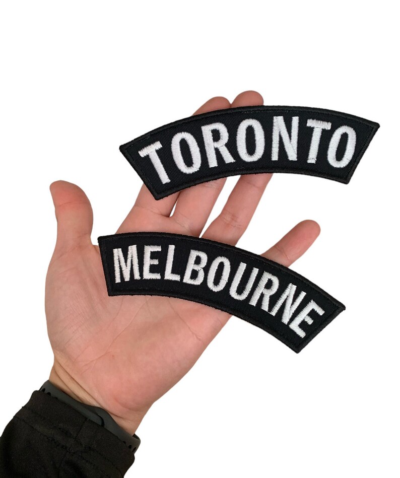 May include: Two black embroidered patches with white text. The top patch reads "TORONTO" and the bottom patch reads "MELBOURNE". Both patches are curved and have a black border.