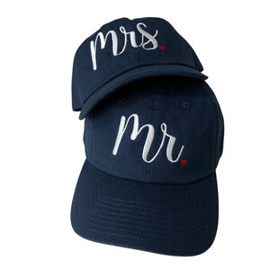 Puede incluir: Dos gorras de béisbol azul marino apiladas una encima de la otra. La gorra superior tiene la palabra "mrs" bordada en escritura blanca con un pequeño corazón rojo. La gorra inferior tiene "mr" bordado en escritura blanca con un pequeño corazón rojo.
