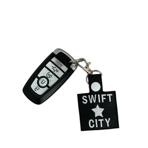 Może przedstawiać: Czarny brelok do kluczy samochodowych ze srebrnym metalowym klipsem przymocowanym do czarnego sk&oacute;rzanego breloka. Brelok ma napis "SWIFT CITY" i białą gwiazdę.