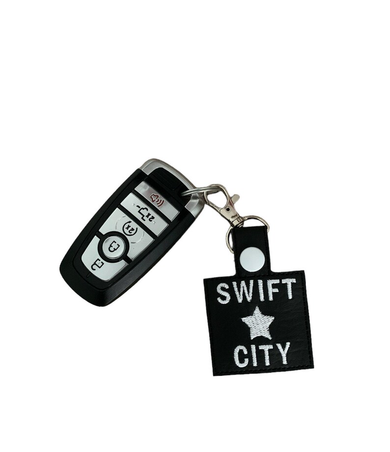 Może przedstawiać: Czarny brelok do kluczy samochodowych ze srebrnym metalowym klipsem przymocowanym do czarnego sk&oacute;rzanego breloka. Brelok ma napis "SWIFT CITY" i białą gwiazdę.