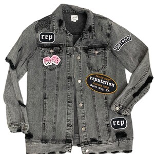 May include: A distressed grey denim jacket with various patches. Patches include the word "rep" in a white oval, a pink dice patch, and a black and gold patch that reads "Taylor Swift Reputation Stadium Tour Swift City, CA".