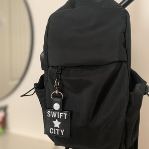 Może przedstawiać: Czarny nylonowy plecak z przednią kieszenią zapinaną na zamek i bocznymi kieszeniami. Do zamka przymocowany jest brelok z napisem "SWIFT CITY" i gwiazdką. Plecak wisi na czarnym metalowym drążku.