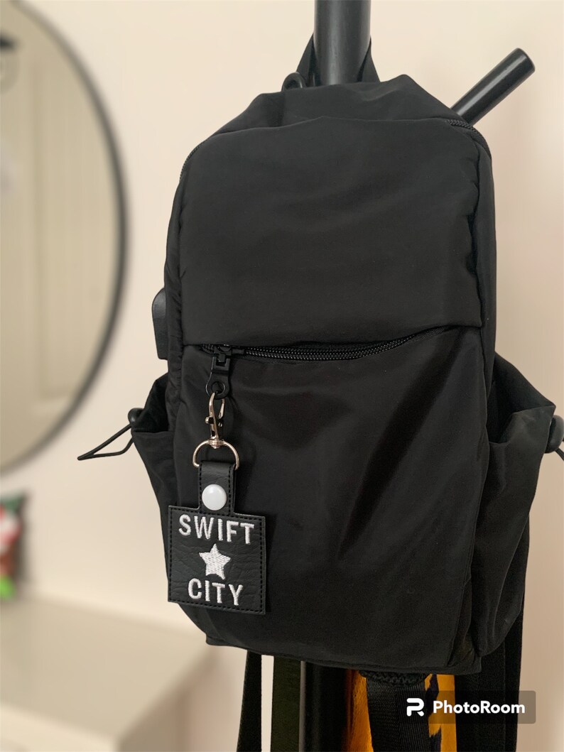Może przedstawiać: Czarny nylonowy plecak z przednią kieszenią zapinaną na zamek i bocznymi kieszeniami. Do zamka przymocowany jest brelok z napisem "SWIFT CITY" i gwiazdką. Plecak wisi na czarnym metalowym drążku.