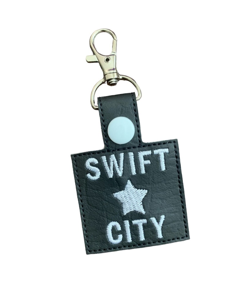 Może przedstawiać: Czarny brelok do kluczy z białym haftem "SWIFT CITY" i gwiazdą. Posiada srebrne zapięcie i jasnoniebieski guzik. Przeznaczony do mocowania do kluczy lub toreb.
