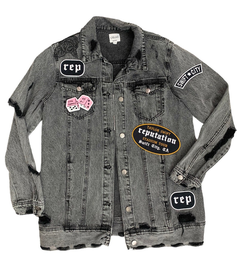 May include: A distressed grey denim jacket with patches. Patches include the word "rep" in a white oval, a pink dice patch, and a black and gold patch that reads "Taylor Swift Reputation Stadium Tour Swift City, CA".
