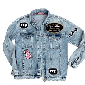 May include: Light blue denim jacket with various patches. Patches include the word "rep" in a black oval, pink dice, and a black oval patch that reads "Taylor Swift Reputation Stadium Tour Swift City, CA" and "Swift City".