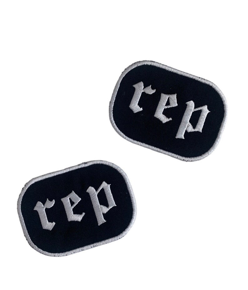 May include: Two black embroidered patches with the word "rep" in white gothic lettering. Each patch has a rounded rectangular shape with a white border. The patches are displayed against a white background.
