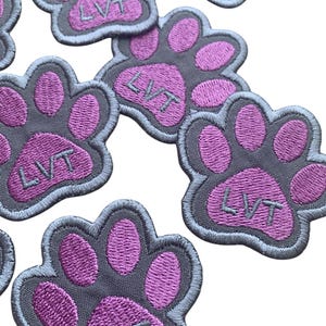 May include: Embroidered paw print patches in shades of gray and purple. Each patch features the letters "LVT" in the center. The patches are designed with a gray border and a white background.