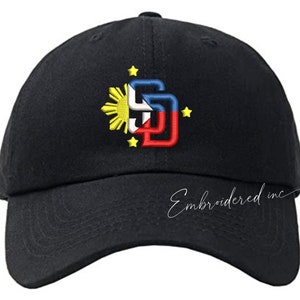 Peut inclure: Casquette de baseball noire avec un motif brodé représentant le soleil et les étoiles du drapeau philippin, ainsi que les lettres "SD" en rouge, blanc et bleu. Le texte "Embroidered inc" est sur le côté.