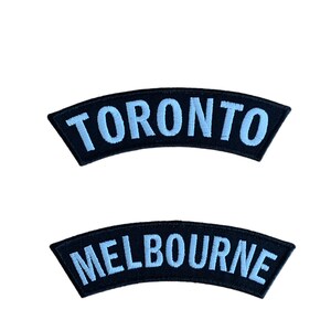 May include: Two black, curved patches with light blue embroidered text. The top patch says "TORONTO", and the bottom patch says "MELBOURNE". These patches are likely for clothing or accessories.
