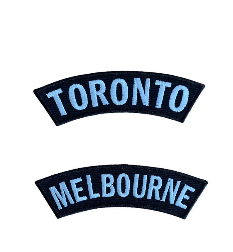 May include: Two black, curved patches with light blue embroidered text. The top patch says "TORONTO", and the bottom patch says "MELBOURNE". These patches are likely for clothing or accessories.