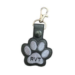 May include: A black paw print keychain with silver hardware. The paw print is embroidered with gray thread and the letters "RVT". The keychain has a silver clasp for easy attachment to keys or bags.
