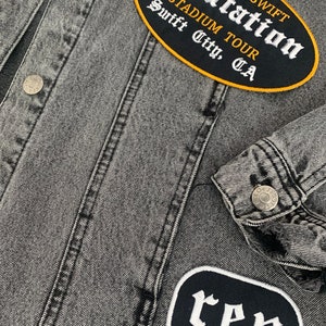 May include: A close-up of a grey denim jacket with silver buttons and stitched seams. The jacket has two embroidered patches: an oval one with "Taylor Swift Reputation Stadium Tour Swift City, CA" and another with the letters "rep".