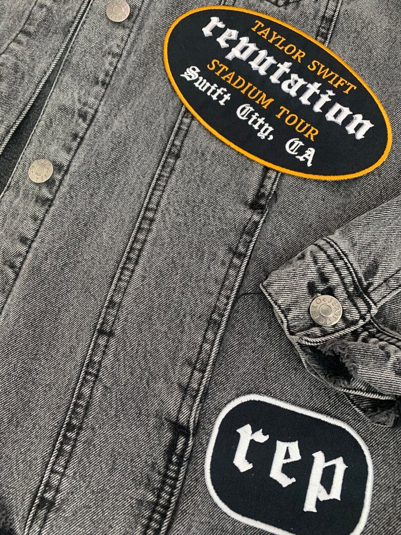 May include: A close-up of a grey denim jacket with silver buttons and stitched seams. The jacket has two embroidered patches: an oval one with "Taylor Swift Reputation Stadium Tour Swift City, CA" and another with the letters "rep".