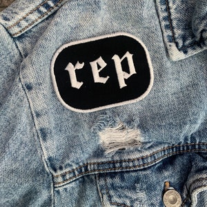 May include: Pale blue denim jacket with distressed detailing. A black patch with the word "rep" in white gothic lettering is sewn on the front. The jacket has a button closure and two front pockets.