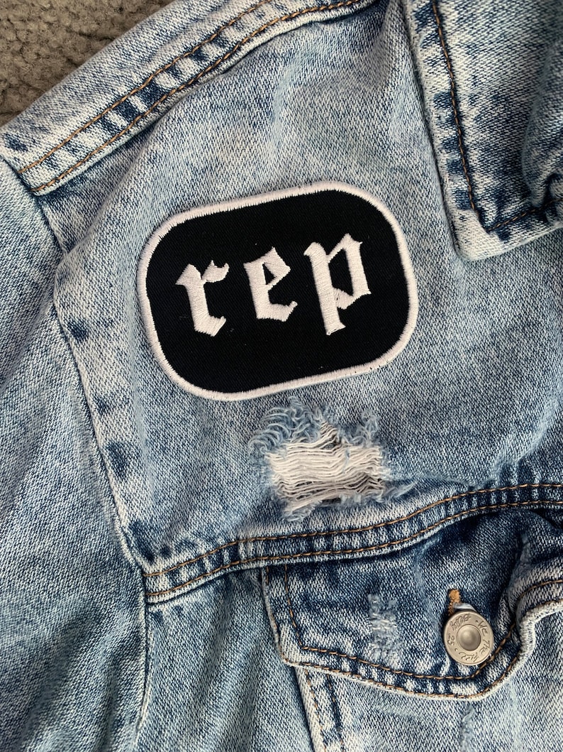 May include: Pale blue denim jacket with distressed detailing. A black patch with the word "rep" in white gothic lettering is sewn on the front. The jacket has a button closure and two front pockets.
