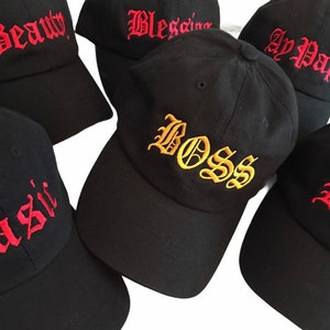 May include: Black baseball caps with embroidered text in various styles. The caps feature words like "Beauty," "Blessing," "Ay Papi," "Boss," and "Basic" in red and gold thread.
