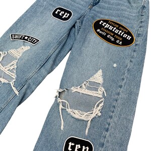 May include: Light blue denim jeans with distressed details and patches. Patches include the text "SWIFT CITY", "rep", "Little Games", "T.S.", and "TAYLOR SWIFT reputation STADIUM TOUR Swift City, CA".