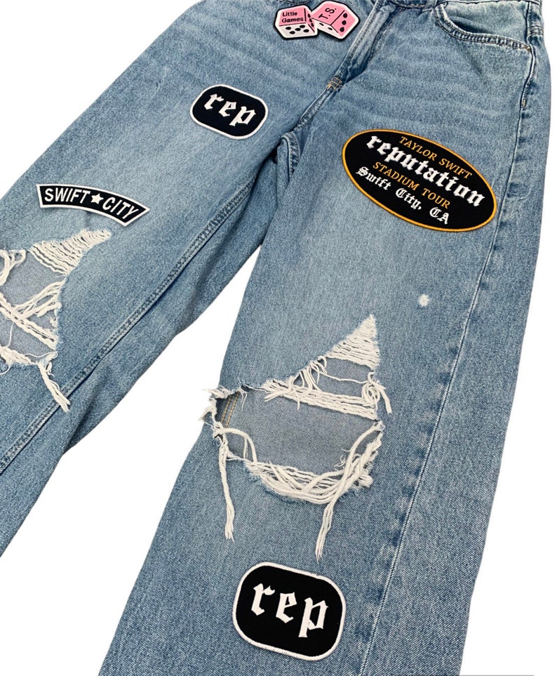 May include: Light blue denim jeans with distressed details and patches. Patches include the text "SWIFT CITY", "rep", "Little Games", "T.S.", and "TAYLOR SWIFT reputation STADIUM TOUR Swift City, CA".