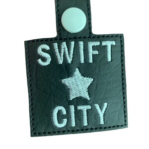 Może przedstawiać: Czarny prostokątny brelok do kluczy z białym haftem "SWIFT CITY" oraz białą gwiazdą. Brelok posiada srebrny metalowy pierścień i biały guzik. Krawędzie są obszyte czarną nicią.