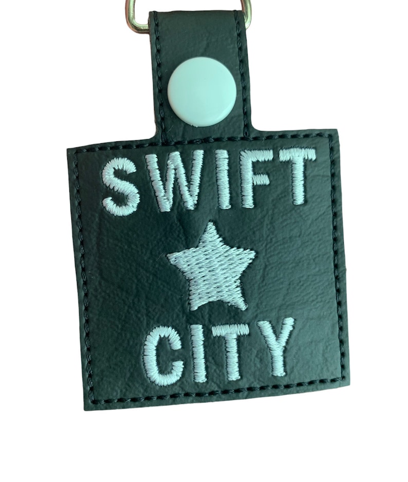 Może przedstawiać: Czarny prostokątny brelok do kluczy z białym haftem "SWIFT CITY" oraz białą gwiazdą. Brelok posiada srebrny metalowy pierścień i biały guzik. Krawędzie są obszyte czarną nicią.