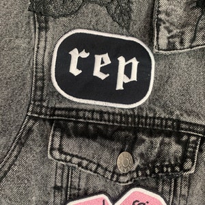 May include: A close-up of a grey denim jacket with black rose embroidery on the shoulder. The jacket has patches with the word "rep" and a pink dice design with the text "Little Games".