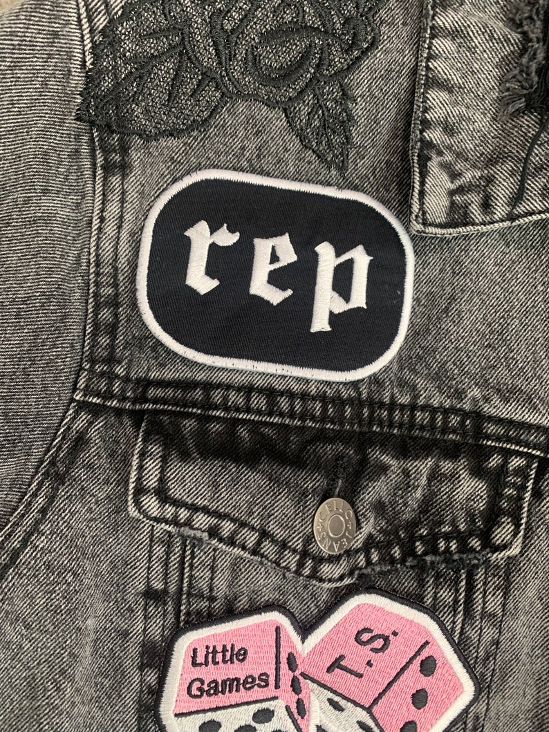 May include: A close-up of a grey denim jacket with black rose embroidery on the shoulder. The jacket has patches with the word "rep" and a pink dice design with the text "Little Games".
