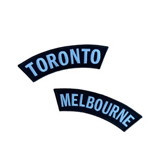 May include: Two embroidered patches. The top patch reads "TORONTO" in light blue letters on a black curved background. The bottom patch reads "MELBOURNE" in light blue letters on a black curved background.