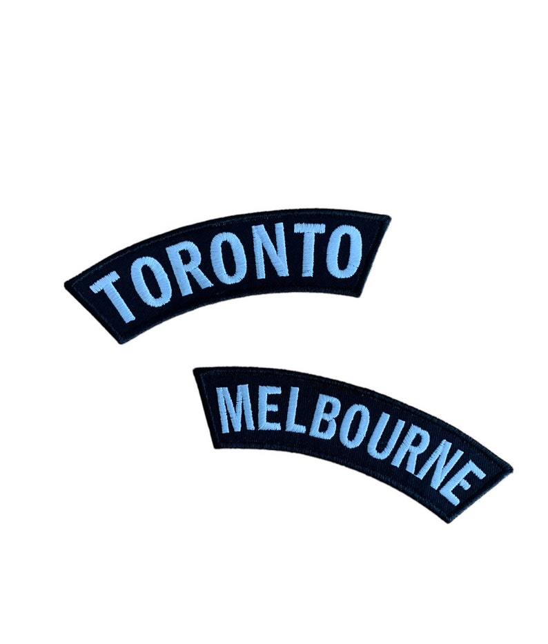 May include: Two embroidered patches. The top patch reads "TORONTO" in light blue letters on a black curved background. The bottom patch reads "MELBOURNE" in light blue letters on a black curved background.