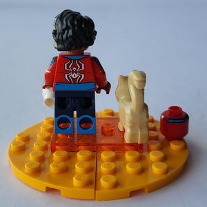 Puede incluir: Una minifigura de Lego con una chaqueta roja y azul con un emblema de ara&ntilde;a blanca, sobre una base amarilla. Una figura de camello beige y un recipiente rojo con una raya azul tambi&eacute;n est&aacute;n en la base. La base est&aacute; hecha de ladrillos Lego amarillos.