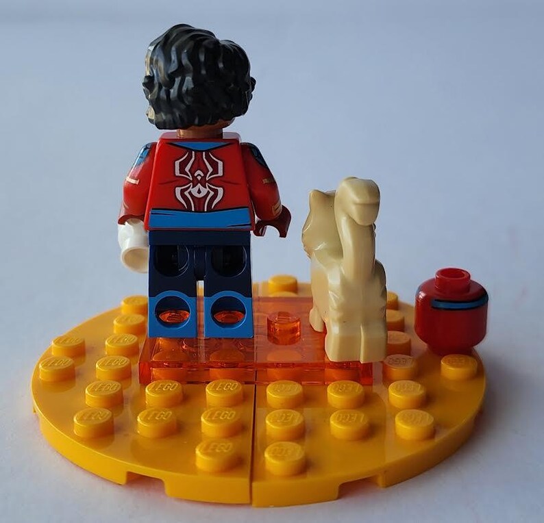 Puede incluir: Una minifigura de Lego con una chaqueta roja y azul con un emblema de ara&ntilde;a blanca, sobre una base amarilla. Una figura de camello beige y un recipiente rojo con una raya azul tambi&eacute;n est&aacute;n en la base. La base est&aacute; hecha de ladrillos Lego amarillos.