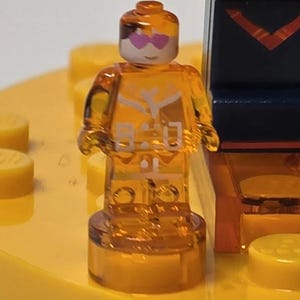 Puede incluir: Una minifigura de Lego naranja transl&uacute;cida con una cara blanca y gafas de sol rosas en forma de coraz&oacute;n. La figura tiene detalles blancos en el torso y est&aacute; de pie sobre una base amarilla. El fondo incluye otras piezas de Lego.