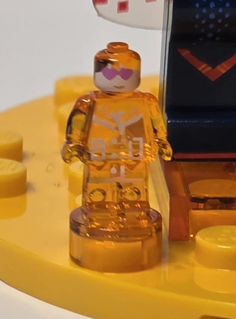 Puede incluir: Una minifigura de Lego naranja transl&uacute;cida con una cara blanca y gafas de sol rosas en forma de coraz&oacute;n. La figura tiene detalles blancos en el torso y est&aacute; de pie sobre una base amarilla. El fondo incluye otras piezas de Lego.