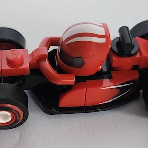 May include: A red and black Lego Ferrari Formula 1 race car with a white stripe on the driver's helmet. The car has the word "Ferrari" printed on the back.