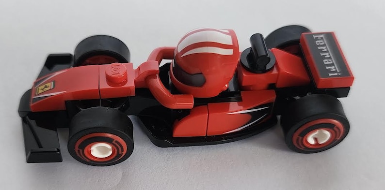 May include: A red and black Lego Ferrari Formula 1 race car with a white stripe on the driver's helmet. The car has the word "Ferrari" printed on the back.