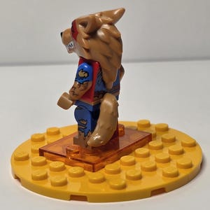 May include: A Lego minifigure of a werewolf with brown fur, red and blue clothing, and a tan tail. The figure stands on an orange rectangular base, which is on a yellow circular base.