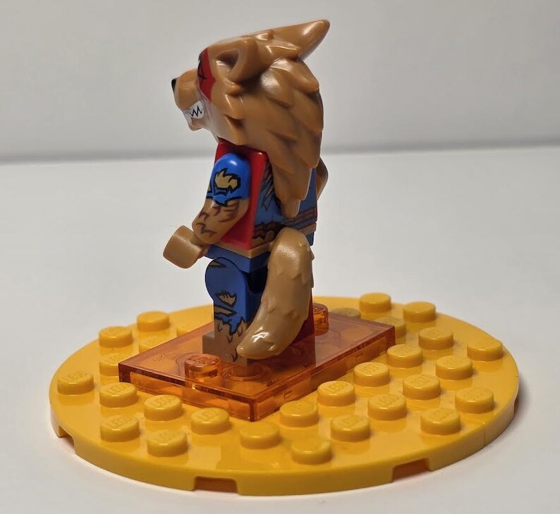 May include: A Lego minifigure of a werewolf with brown fur, red and blue clothing, and a tan tail. The figure stands on an orange rectangular base, which is on a yellow circular base.
