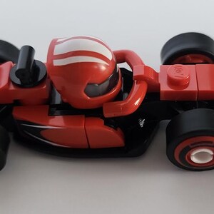 May include: A red and black Lego Ferrari Formula One race car with a white stripe on the helmet of the driver. The car has the word "Ferrari" printed on the side.