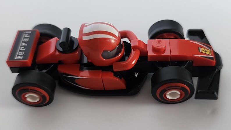 May include: A red and black Lego Ferrari Formula One race car with a white stripe on the helmet of the driver. The car has the word "Ferrari" printed on the side.