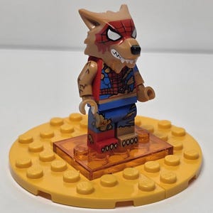 May include: A Lego minifigure of a wolf-like character with a red and blue spider-themed costume. The figure stands on a yellow Lego base with an orange transparent platform. The character has a brown head with white eyes and a red spider web design.