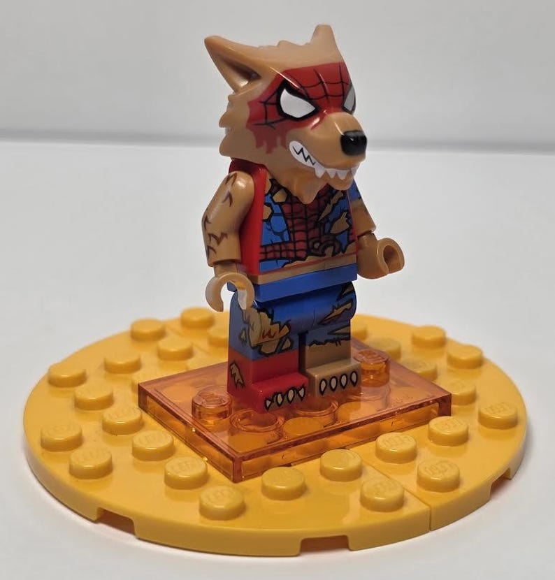 May include: A Lego minifigure of a wolf-like character with a red and blue spider-themed costume. The figure stands on a yellow Lego base with an orange transparent platform. The character has a brown head with white eyes and a red spider web design.
