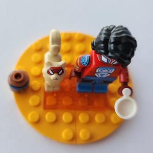 Puede incluir: Una minifigura de Lego Spider-Man con afro negro, traje rojo y azul y un accesorio blanco. Un gato blanco con m&aacute;scara de Spider-Man y un objeto marr&oacute;n y azul est&aacute;n sobre una base amarilla.