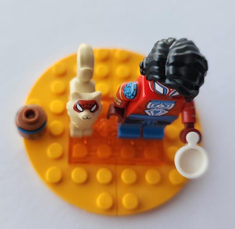 Puede incluir: Una minifigura de Lego Spider-Man con afro negro, traje rojo y azul y un accesorio blanco. Un gato blanco con m&aacute;scara de Spider-Man y un objeto marr&oacute;n y azul est&aacute;n sobre una base amarilla.