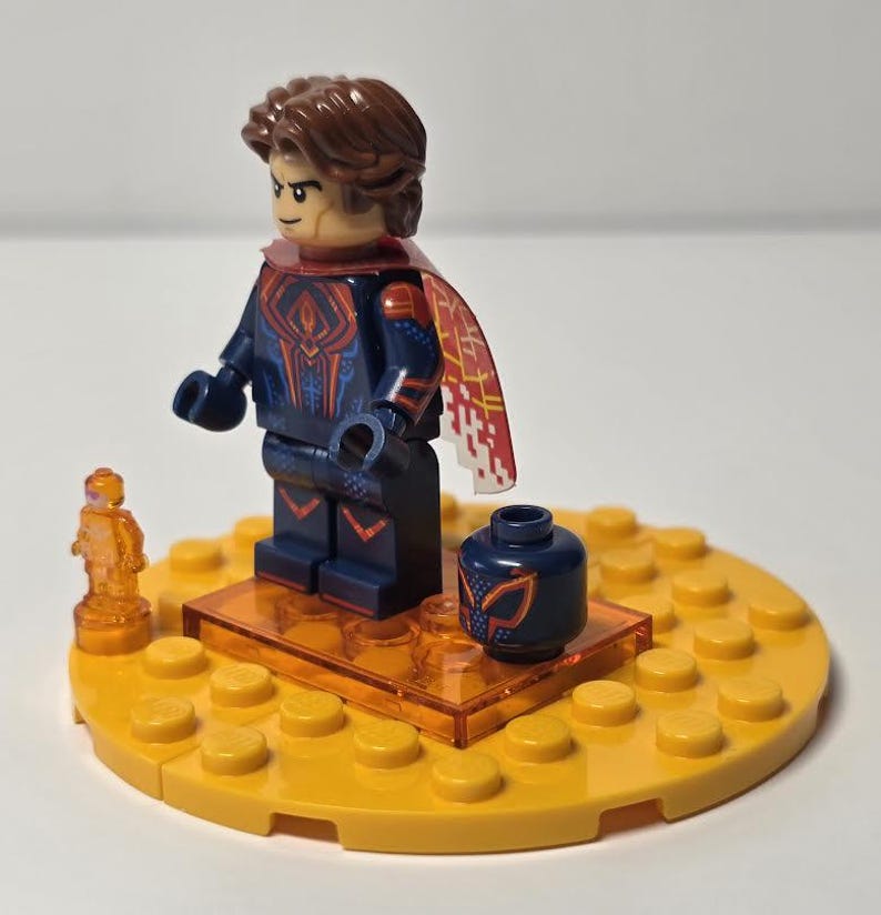 Puede incluir: Una minifigura de Lego de un superh&eacute;roe con cabello casta&ntilde;o, un traje azul oscuro con detalles rojos y dorados, y una capa roja y blanca. La figura est&aacute; sobre una base amarilla con una peque&ntilde;a figura naranja y una cabeza azul.