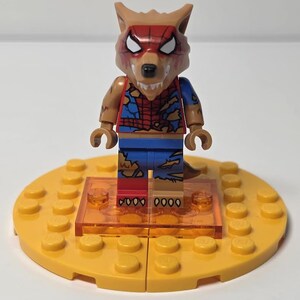 May include: A Lego minifigure of a wolf-like creature with a red and brown mask, brown arms, and a torn blue and red outfit. The figure stands on a yellow Lego base with an orange transparent platform.